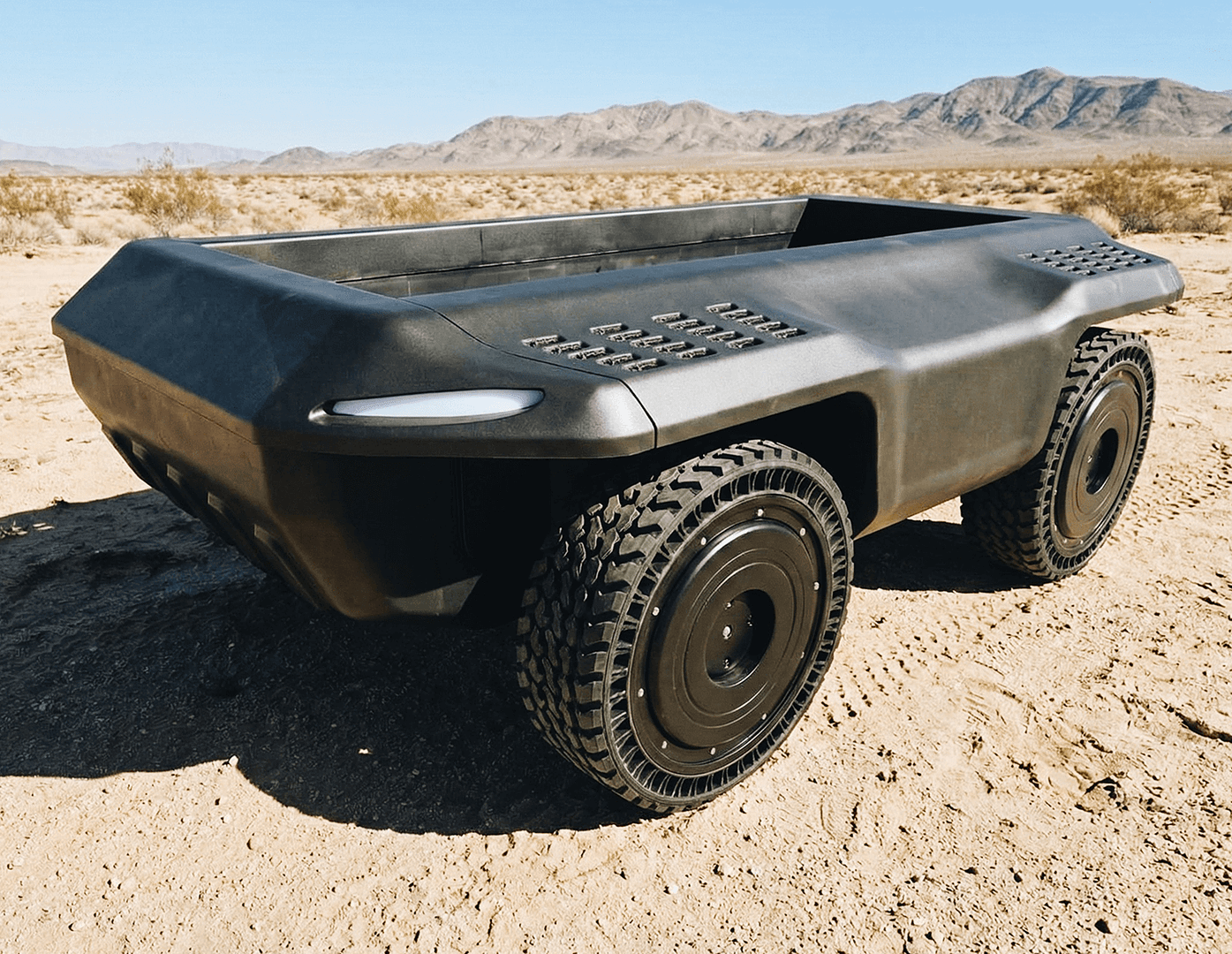 A black, rugged, four-wheeled autonomous vehicle with no visible cabin or controls sits on sandy terrain in a desert landscape with distant mountains under a clear blue sky.