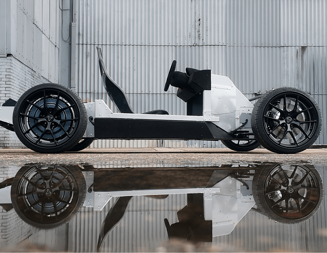 A minimalist, open-frame electric vehicle chassis with exposed wheels and no body panels is parked outside on concrete, reflected in a puddle of water. The background is a corrugated metal building.