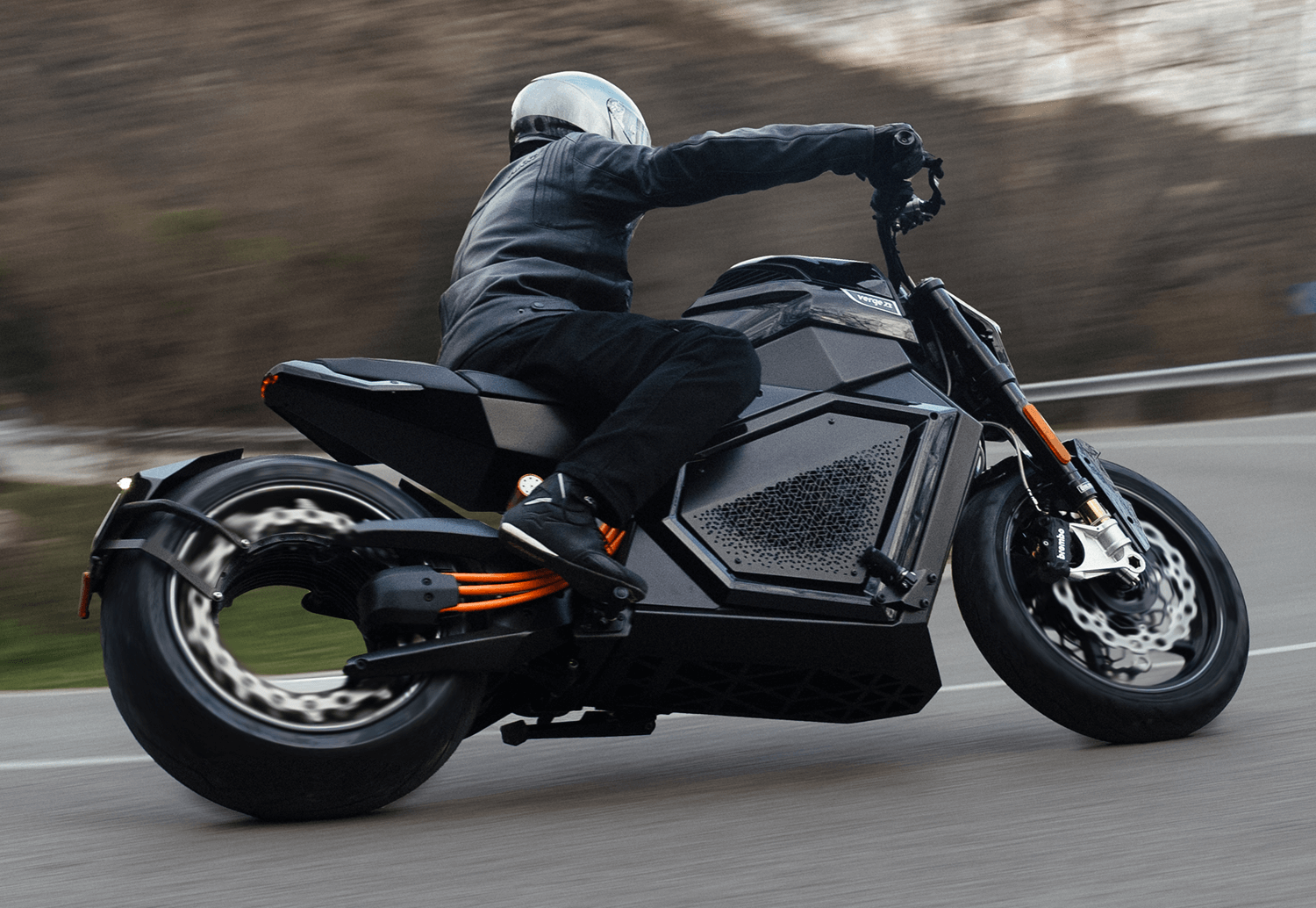 A person wearing a helmet and black leather jacket rides a black electric motorcycle at speed, leaning into a curve on a paved road, with blurred trees and hills in the background.