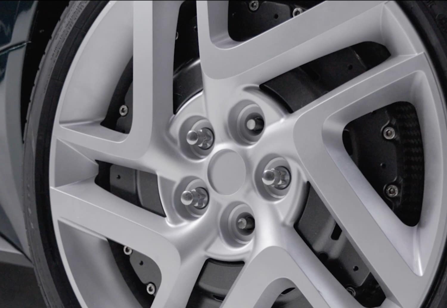 Close-up of a sleek, modern alloy car wheel with a silver five-spoke design. The black tire and brake disc with visible bolts are partially visible behind the wheel.
