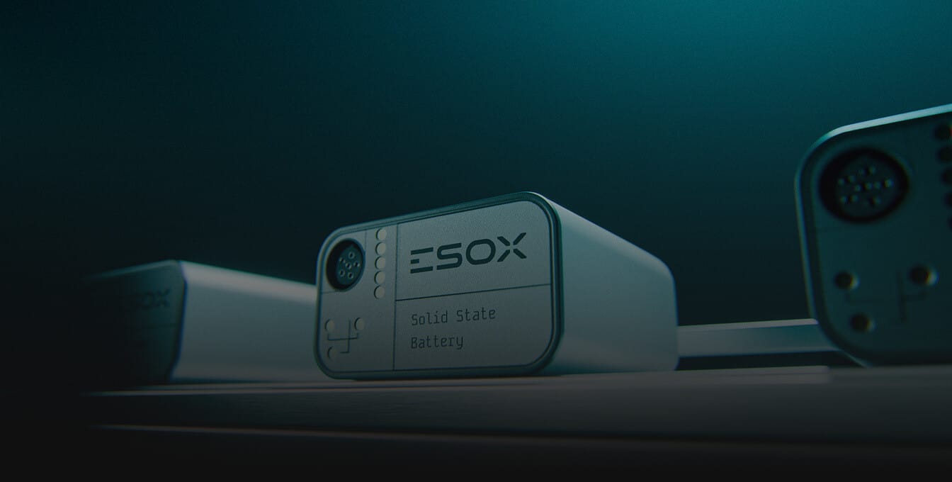 Close-up of a rectangular ESOX solid state battery with a plug connector, illuminated in a dark, teal-lit environment, sitting on a flat surface with part of another device visible beside it.