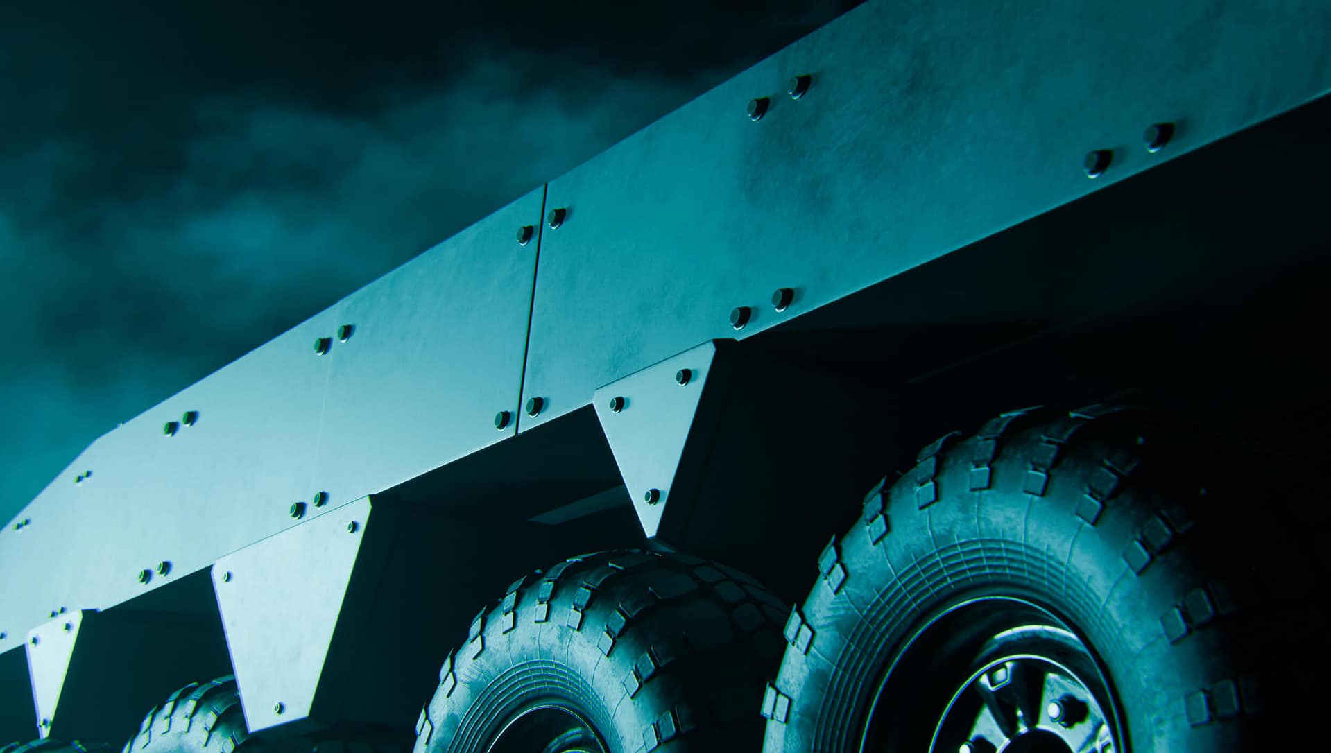 A close-up, low-angle view of a military armored vehicle showing large rugged tires and angular metal plating under moody, teal-tinted lighting against a cloudy night sky.