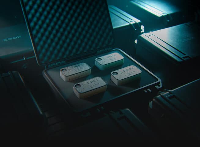 A hard case with foam padding is open to display four small, rectangular electronic devices labeled "CBOX." The case is surrounded by other similar closed cases in a dim, blue-lit environment.