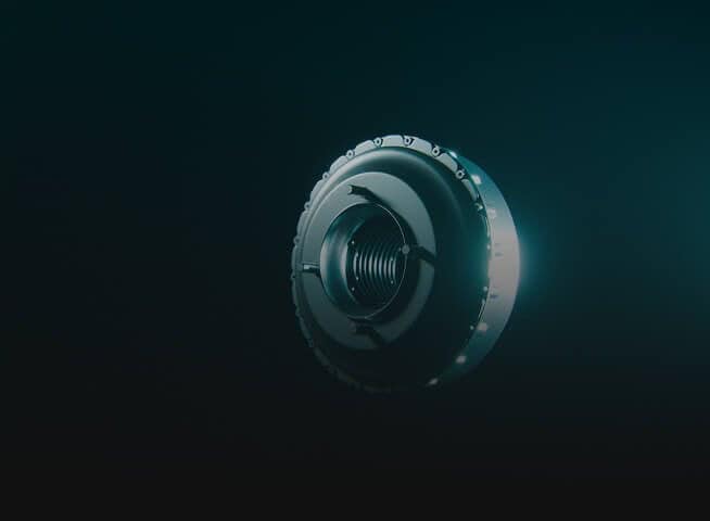 A metallic, circular mechanical part with grooves and a central opening, illuminated against a dark, gradient background. The object appears to be floating and is lit from the right side.