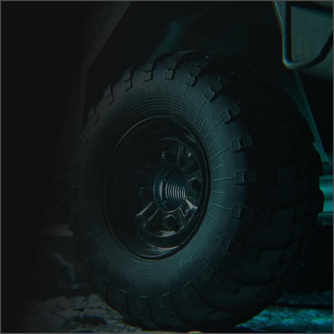 Close-up of a rugged, black off-road vehicle tire with deep tread patterns, mounted on a dark metal wheel, on a rough surface with dim lighting.