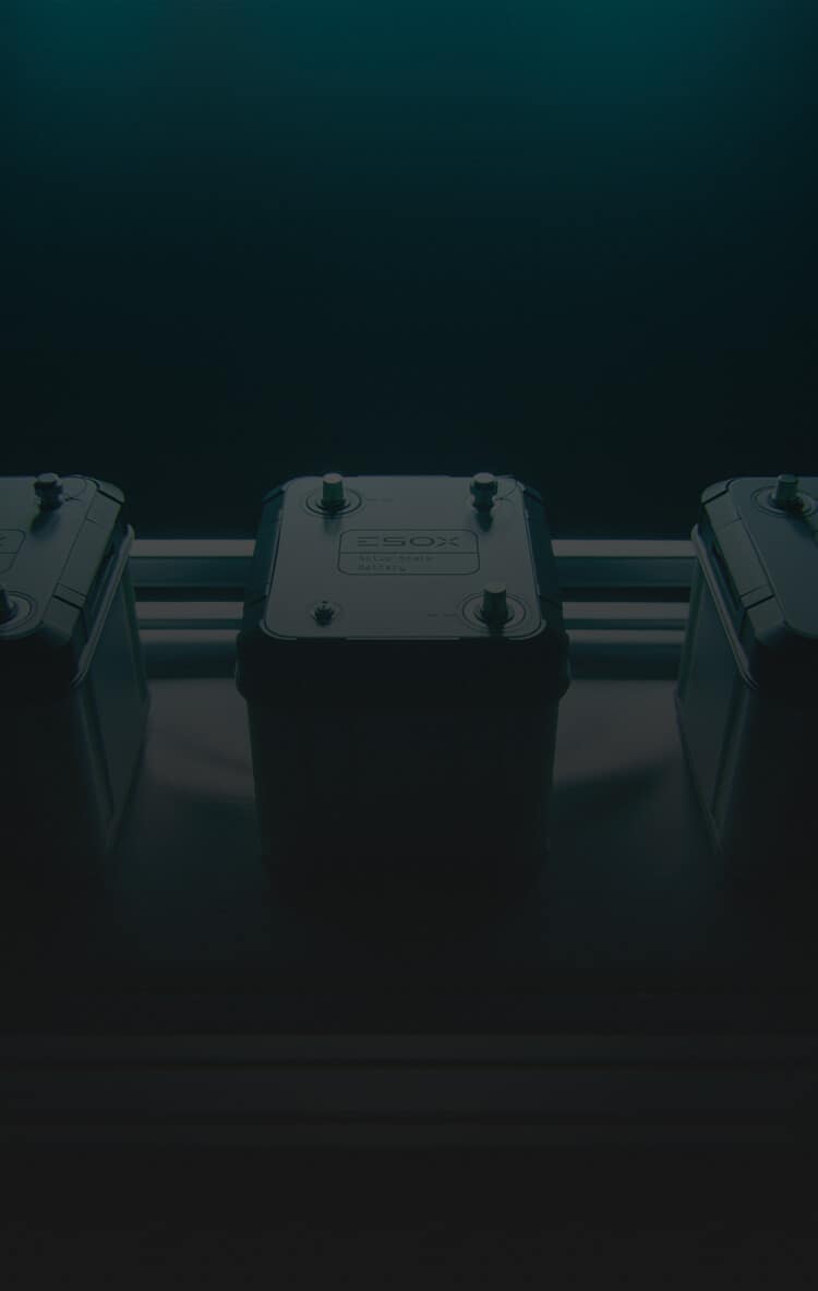 A dark, moody image of a battery pack with two metal terminals on top, placed on a metallic surface, with two similar objects partially visible on either side.