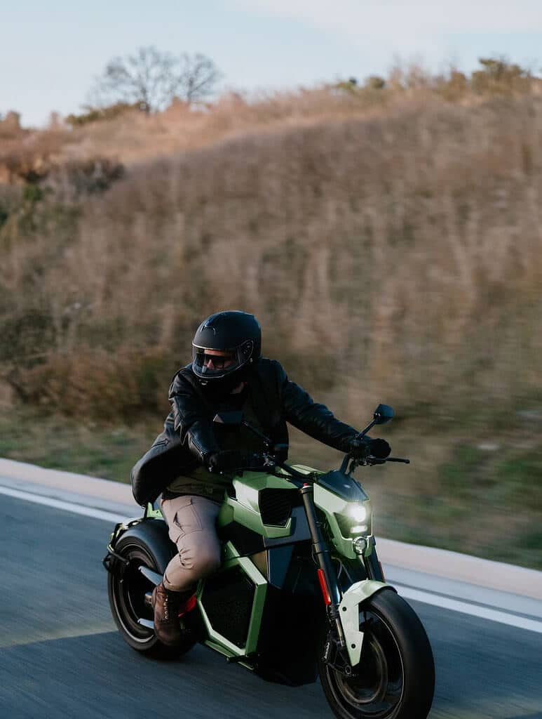 A close-up of a man driving an electric motorcycle. The driver wears a helmet, a black leather jacket and cargo pants. The motorcycle lights are on, as it is at twilight.