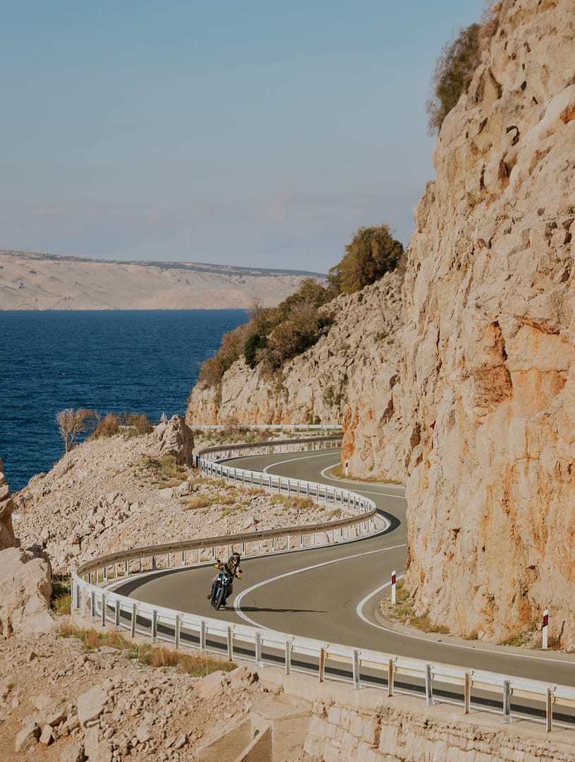 A Verge electric motorcycle rides on a serpentine-like mountain road by the seaside on a sunny day with cliffs alongside the road.