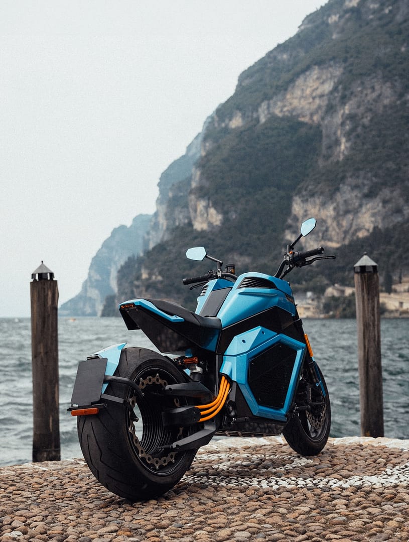 An electric motorcycle parked and captured in the image from the backside, on a pier by the seaside with dark turquoise moving sea and dramatic cliffs at the back
