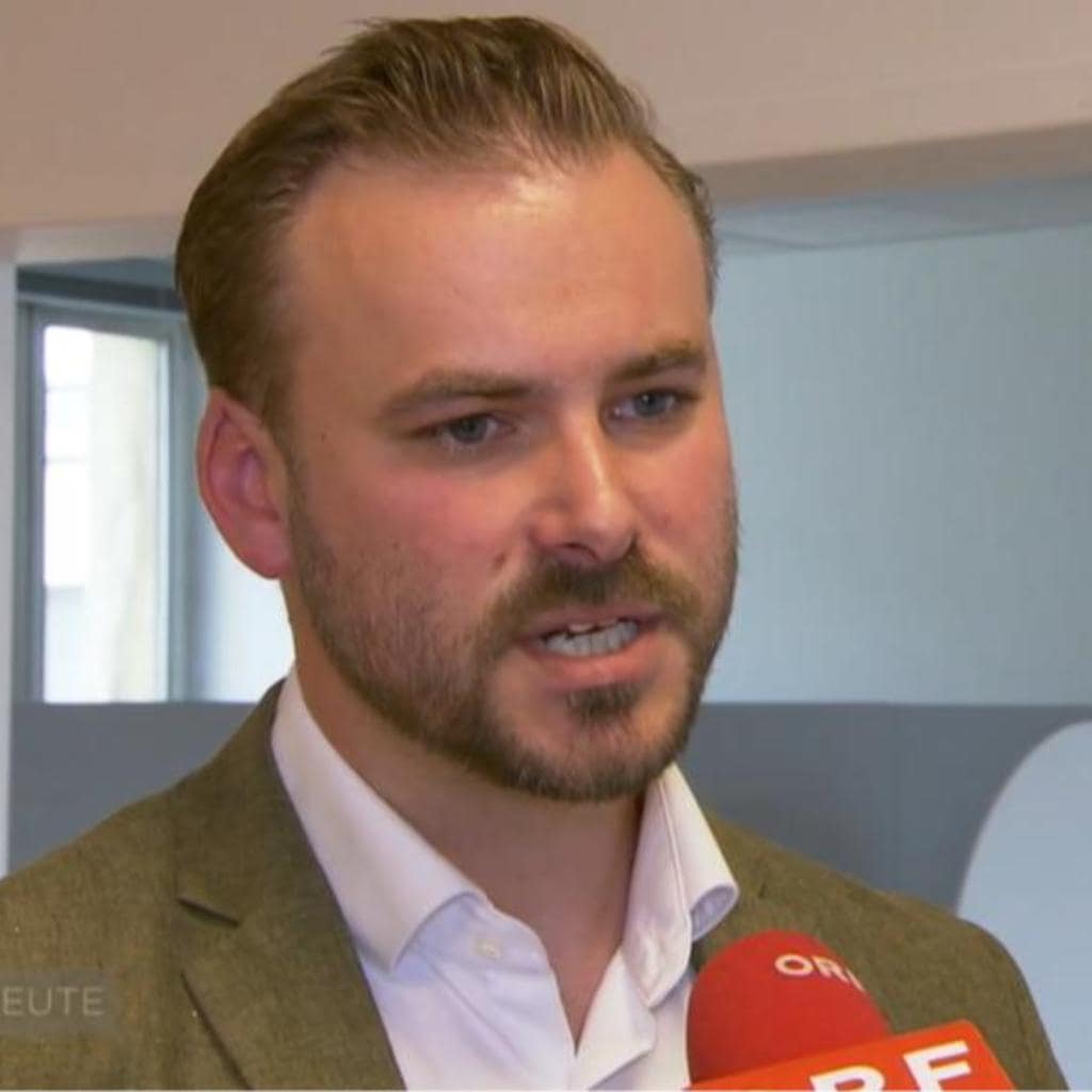 A man with short hair and a beard speaks into a red microphone. He is wearing a white shirt and a green blazer. The background is an indoor setting with light-colored walls.