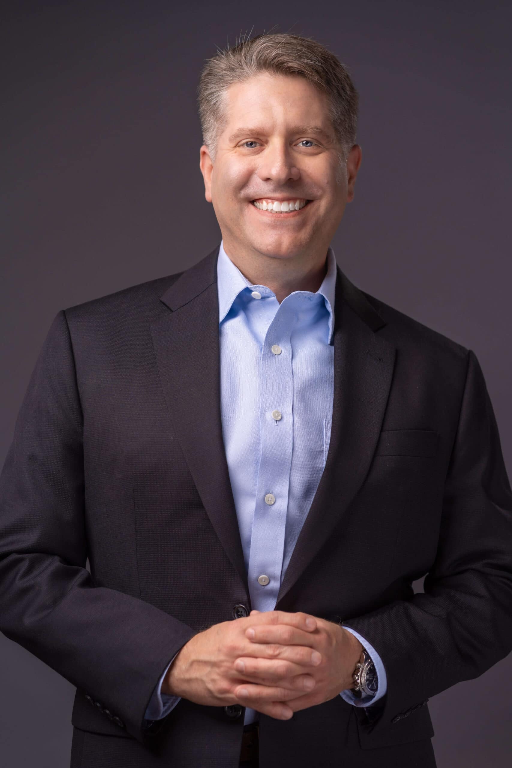 A person in a dark suit and light blue shirt smiles while standing against a gray background. They have short, light-colored hair and are clasping their hands in front.