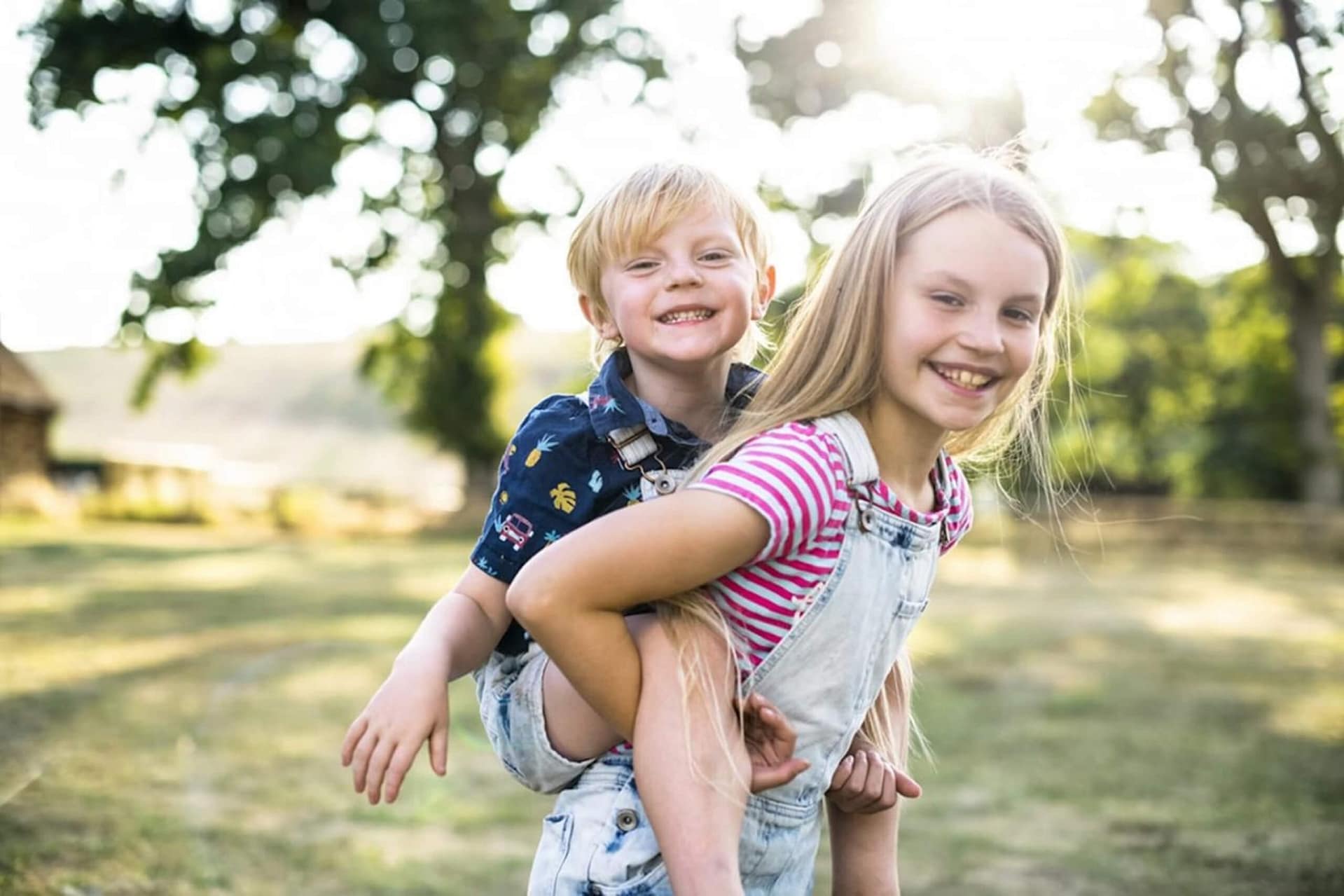 A smiling girl gives a piggyback ride to a laughing younger boy outdoors on a sunny day, with green trees and sunlight in the background.