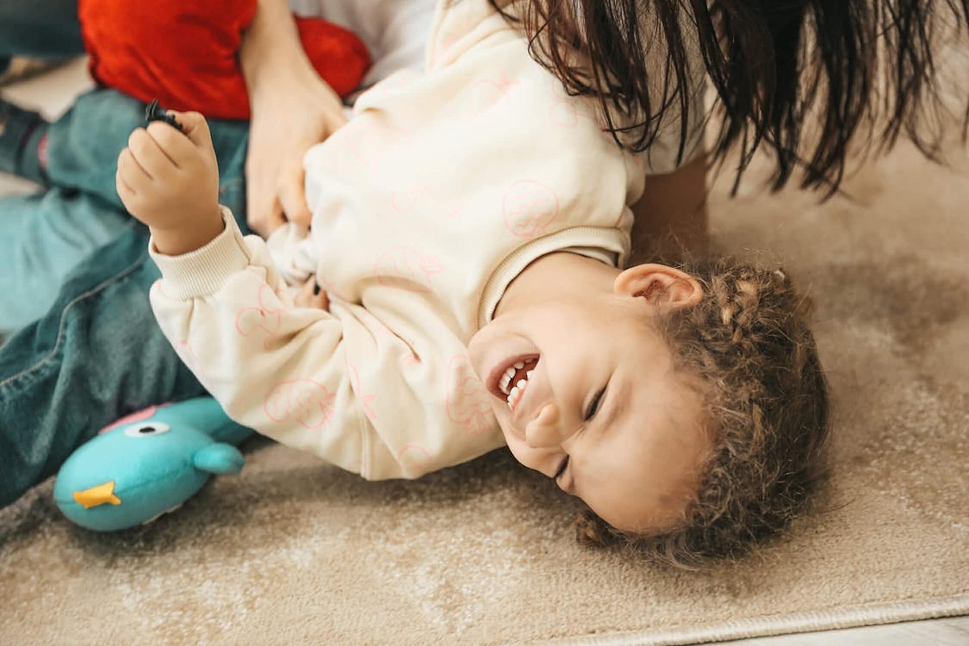 A young child laughs joyfully while lying on a carpeted floor, with a stuffed toy bird nearby and an adult partially visible, playing with them.