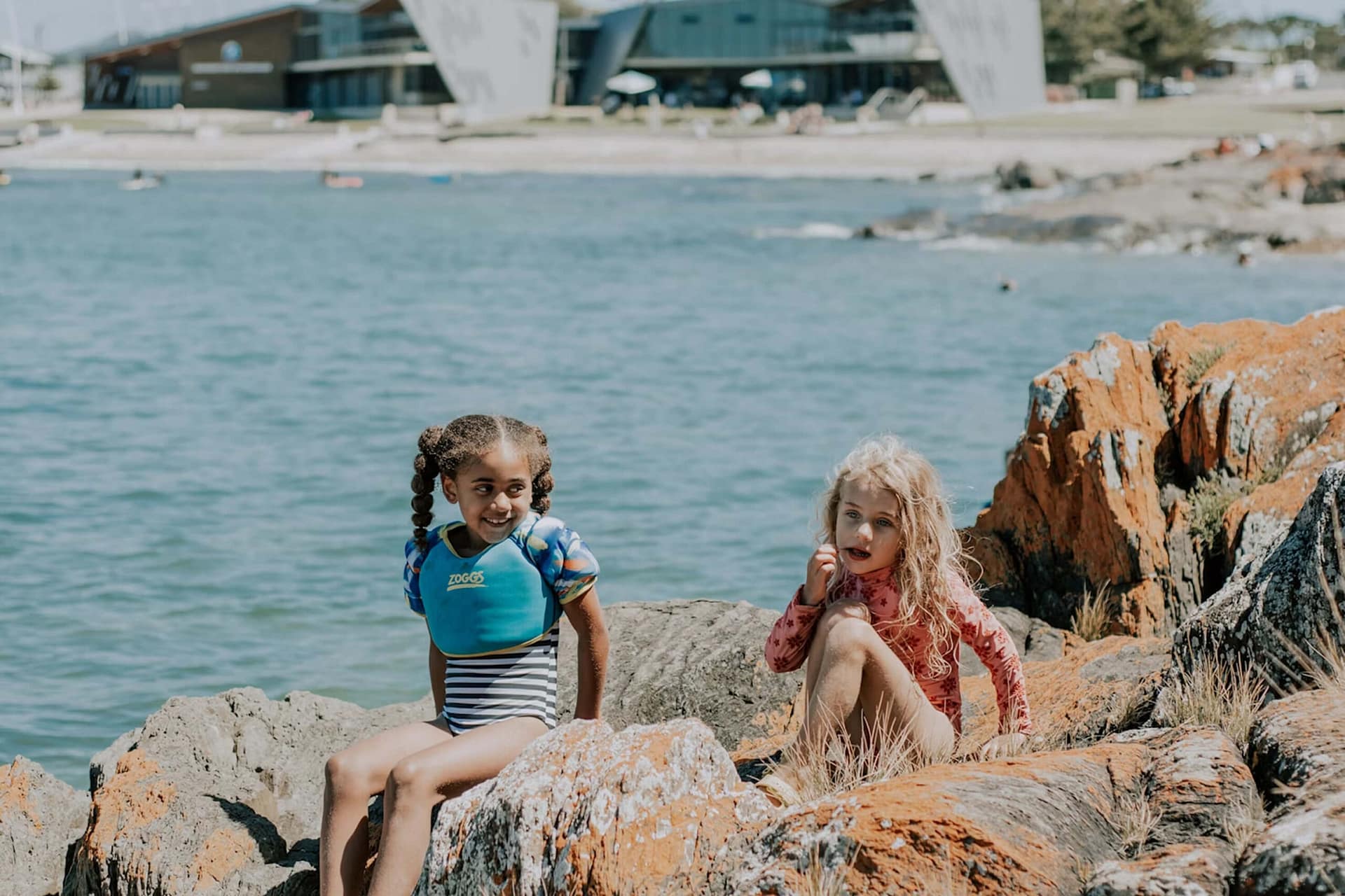 Two young girls in swimsuits sit on orange-lichen-covered rocks by the water. One girl smiles while holding a blue float vest, and the other looks thoughtful. Buildings and a shoreline are visible in the background.