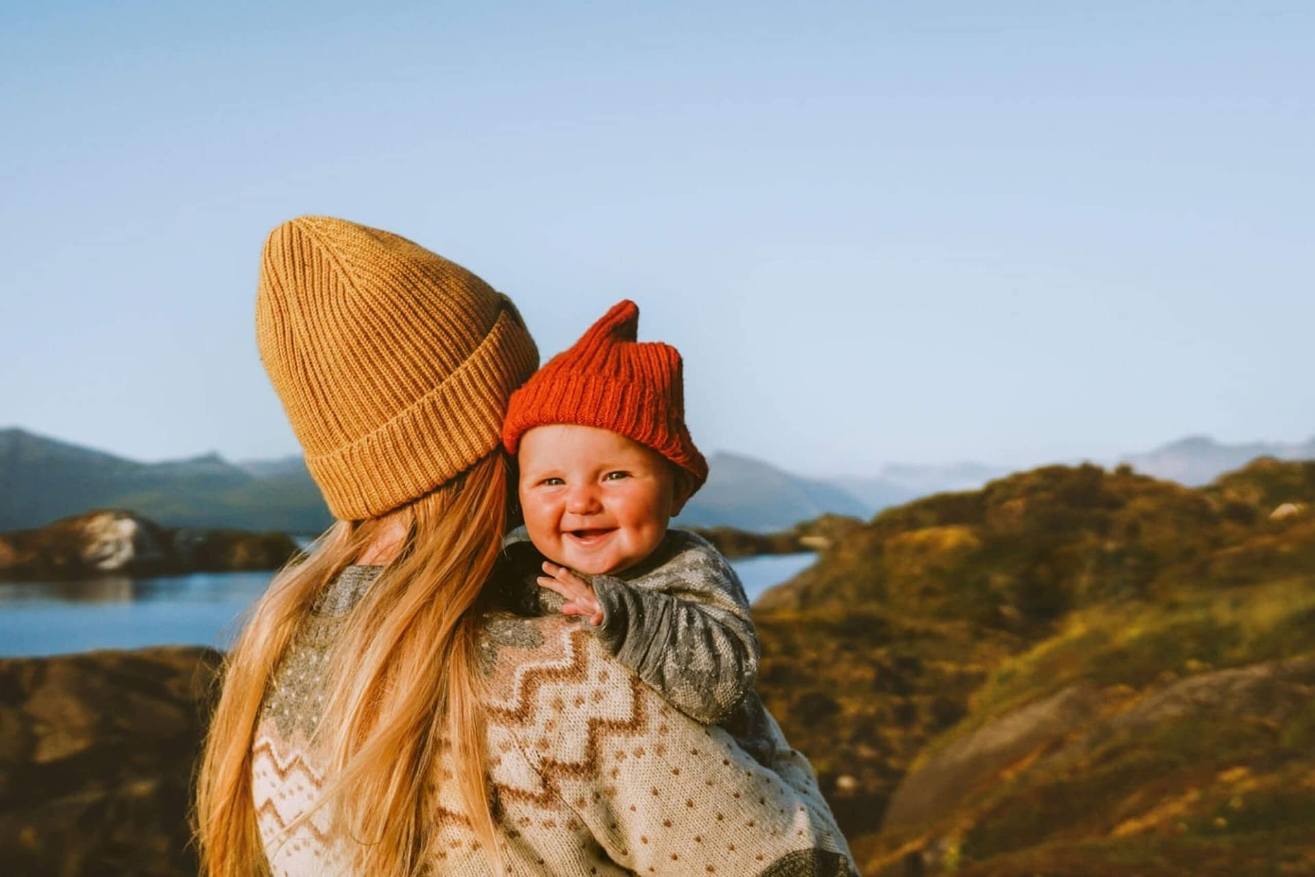 A woman with long blonde hair wearing a tan knit hat holds a smiling baby in a red knit hat, outdoors near a lake and mountains under a clear blue sky.