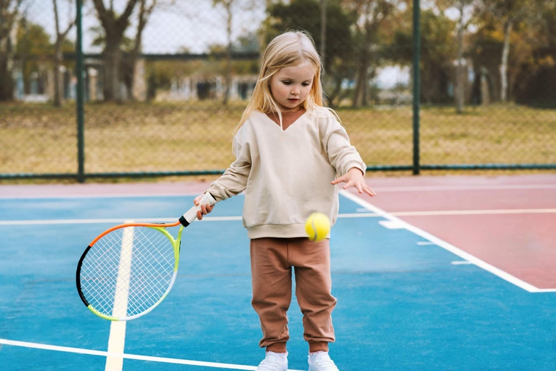 A young girl stands on a blue tennis court, holding a racket in one hand while bouncing a tennis ball with the other. She wears a beige sweater, brown pants, and white sneakers. Trees and a fence are visible in the background.