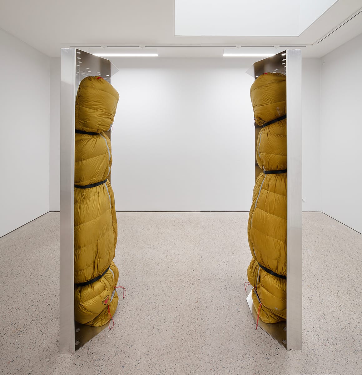 Human-sized figures, wrapped in green thermal sleeping bags and suspended upside down inside stainless-steel trays.