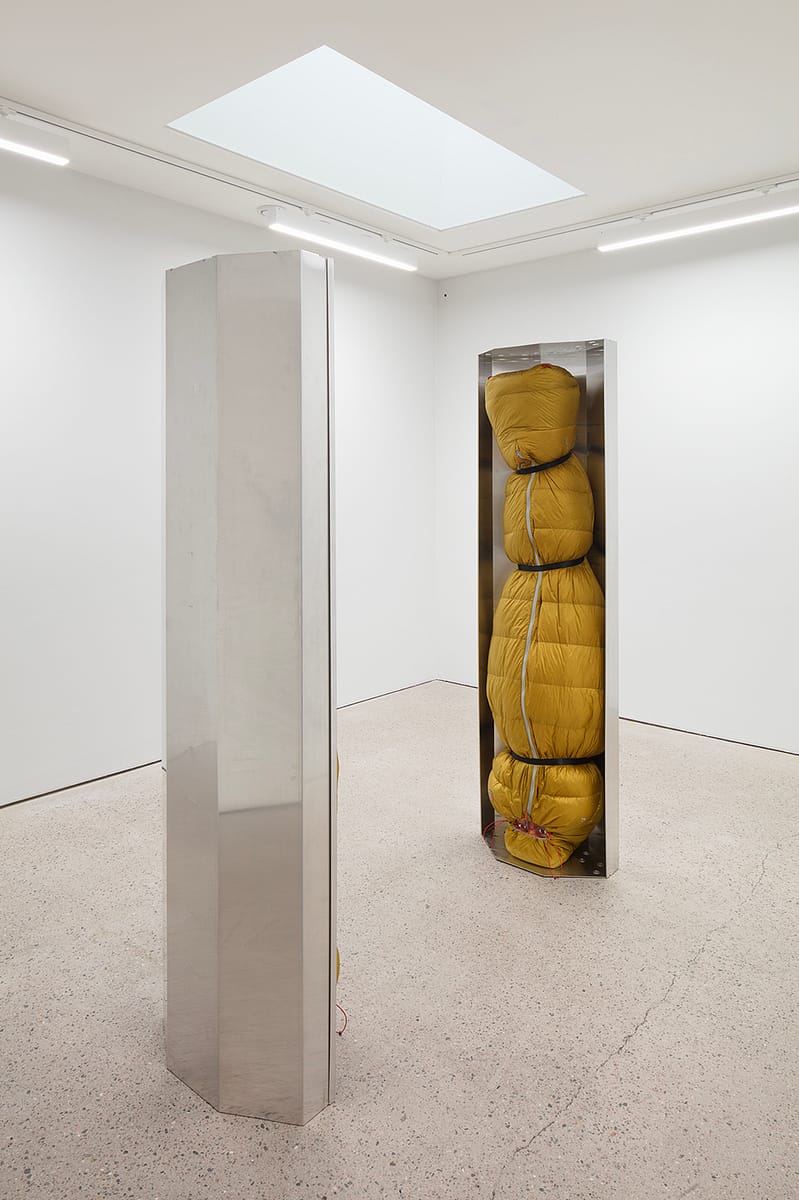 Human-sized figures, wrapped in green thermal sleeping bags and suspended upside down inside stainless-steel trays.