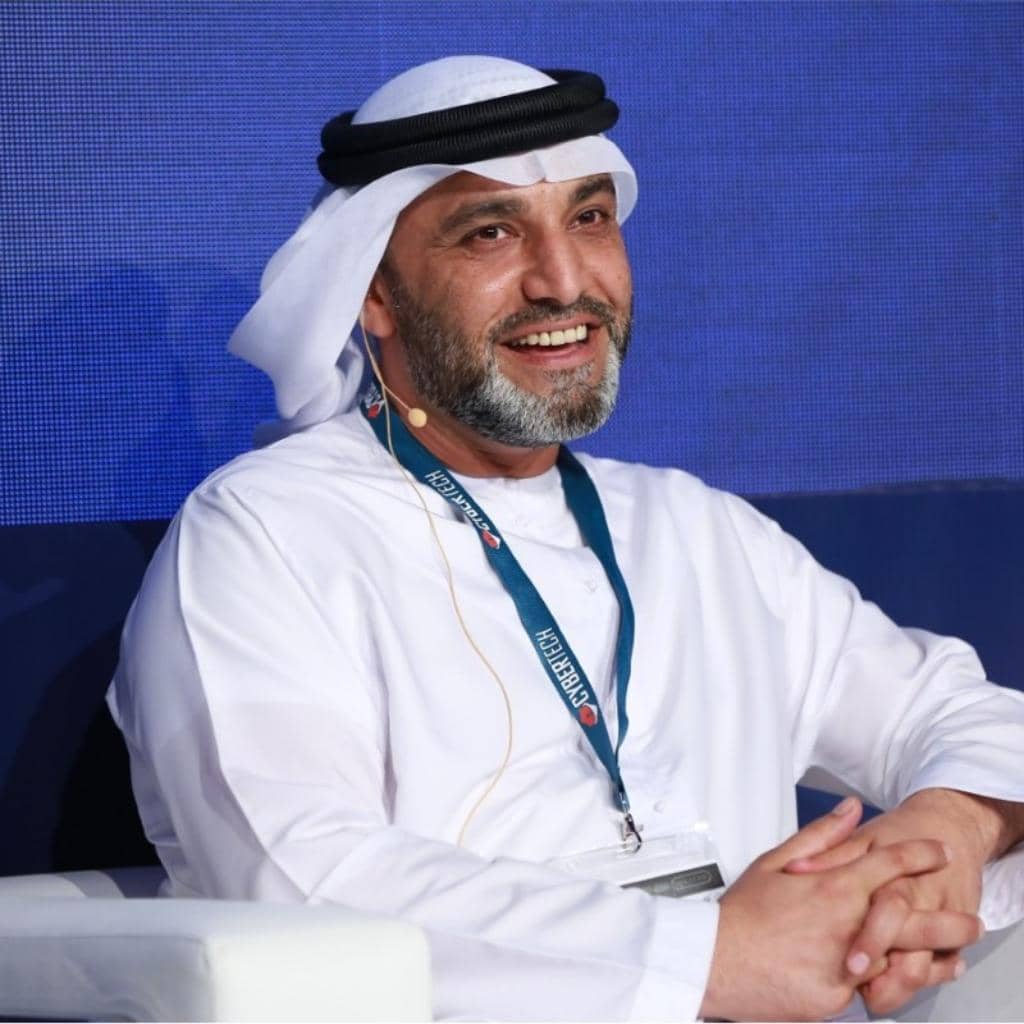 A man wearing a traditional white thobe and headscarf sits smiling in a chair. He is wearing a lanyard and has a microphone headset, suggesting he may be speaking at an event or conference. The background is blue.
