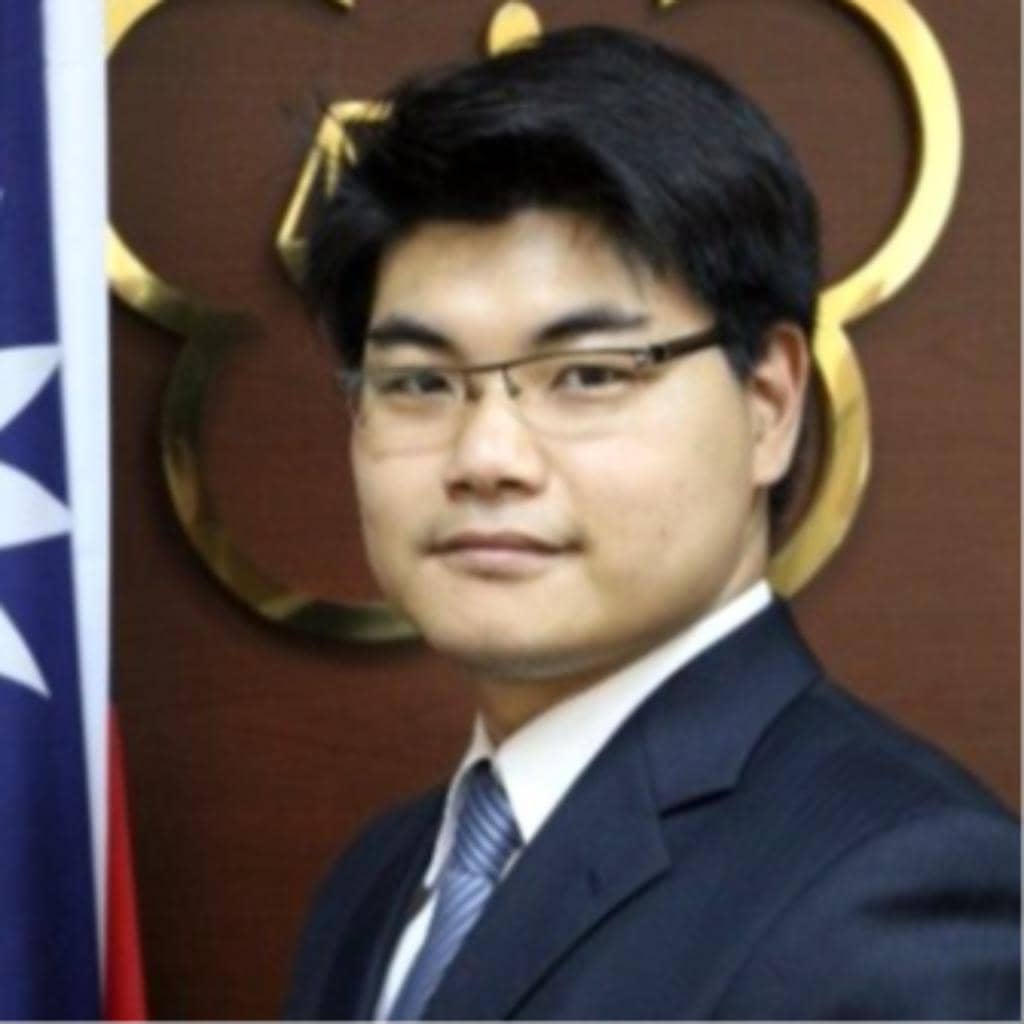 A person wearing glasses and a suit stands in front of a wooden background featuring a stylized emblem. There is a partial view of a flag to the left.