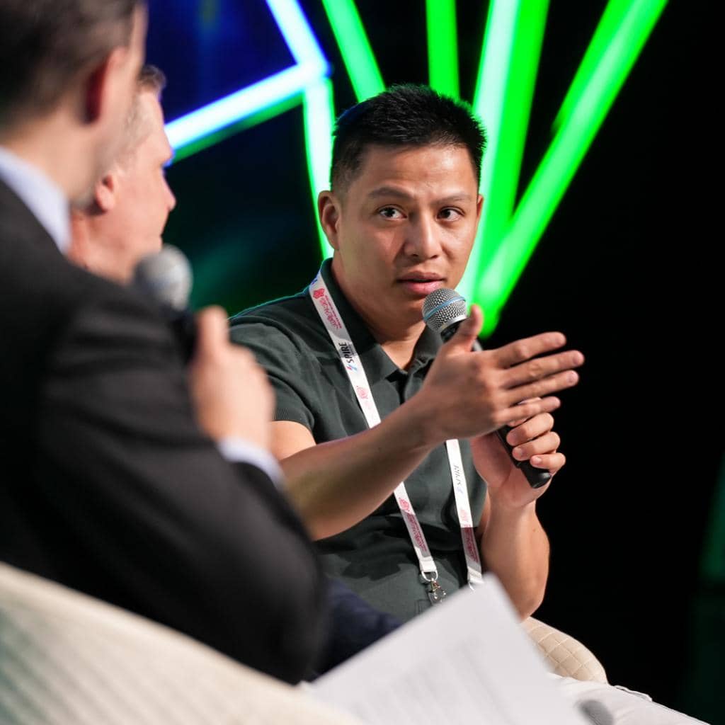 A person speaking into a microphone while gesturing with their hand during a panel discussion. They are seated among other people holding microphones. Bright green lights are in the background.