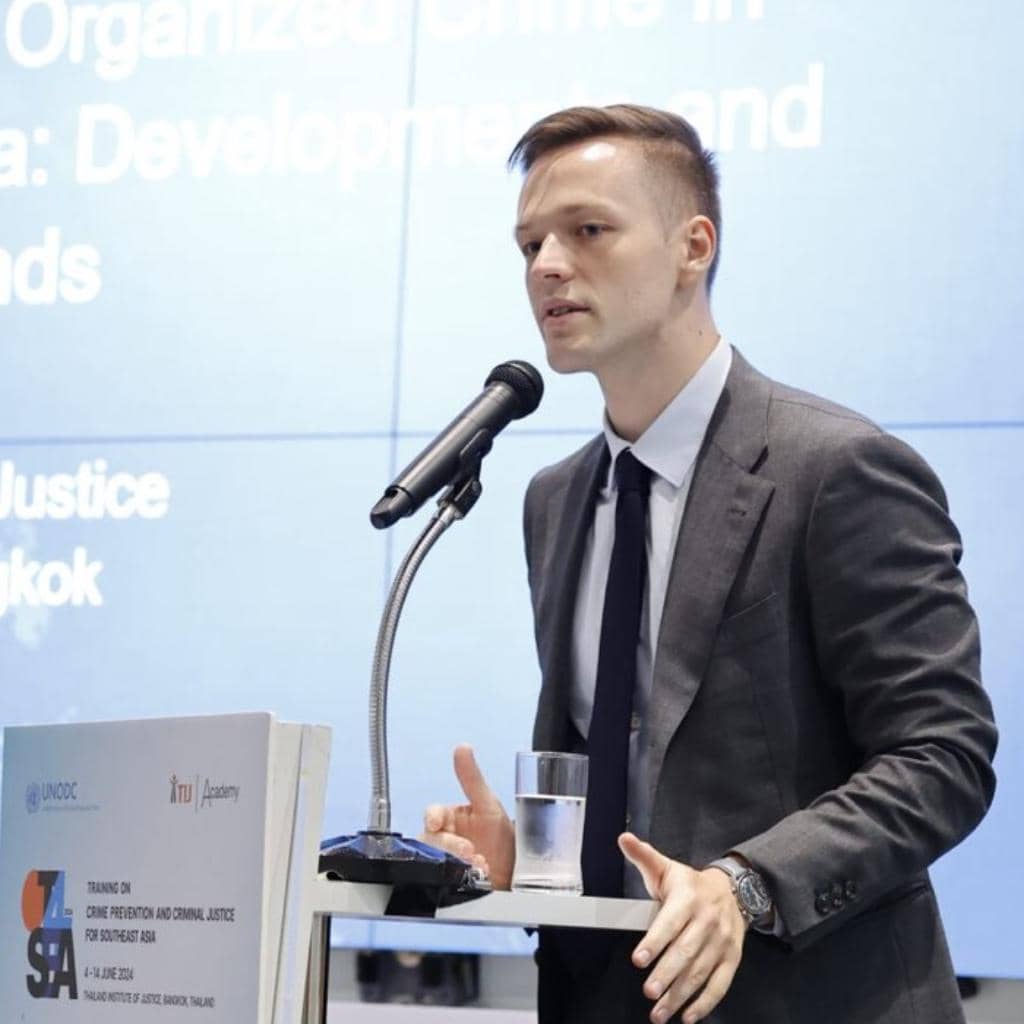 A man in a suit speaks at a podium with a microphone. He holds a glass of water. A screen in the background displays text related to crime prevention and justice. A logo and conference details are on the podium.