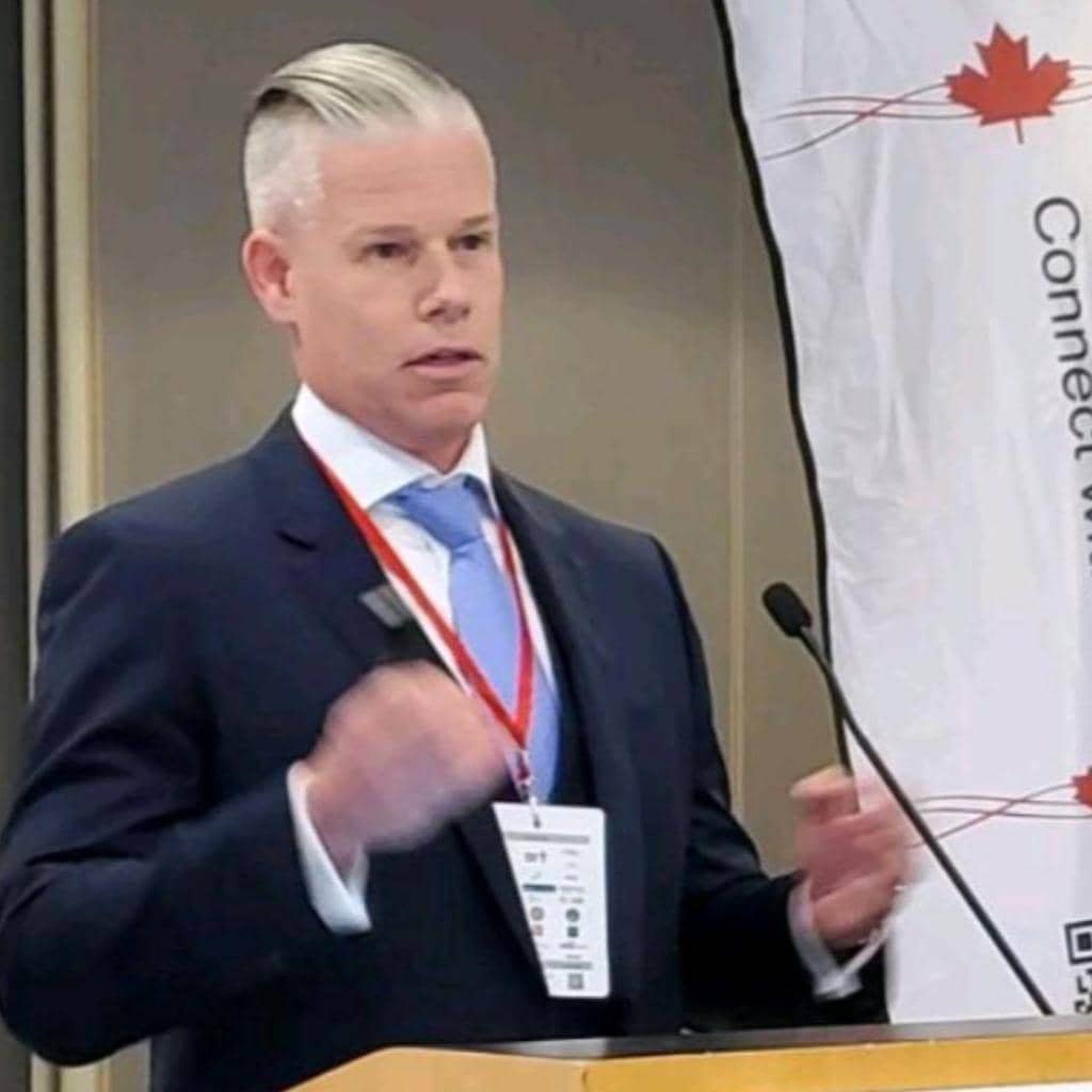 A person in a suit and tie is speaking at a podium. They're holding a microphone and wearing a name badge. The background features a banner with partial text and a maple leaf design.
