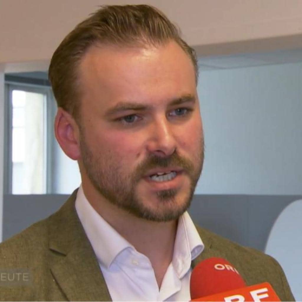 A man with short hair and a beard speaks into a red microphone. He is wearing a white shirt and a green blazer. The background is an indoor setting with light-colored walls.