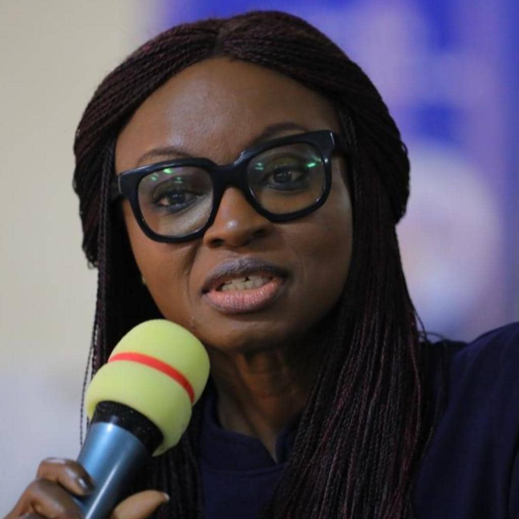 A woman with glasses and braided hair holds a microphone with a green and red band. She appears to be speaking, set against a blurred background.