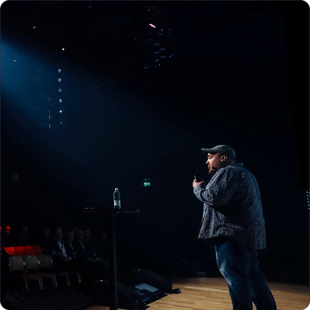 A person wearing a cap and casual clothing is speaking on stage with a microphone. A spotlight shines on them, with an audience partially visible in the dimly lit background. A water bottle is on a nearby table.