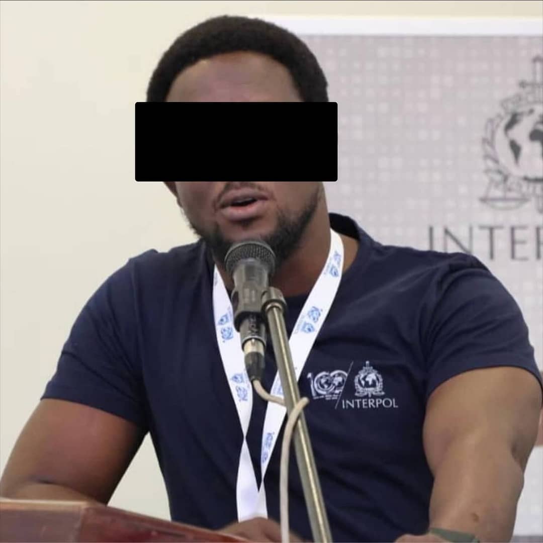 A person wearing a navy blue INTERPOL shirt and a lanyard speaks at a podium with a microphone; their eyes are obscured by a black rectangle for anonymity.