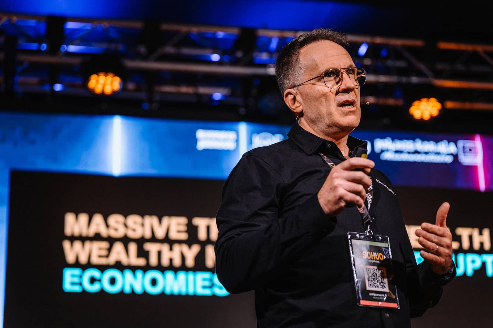 A person is speaking on stage holding a microphone. They're wearing glasses and a black shirt with a conference badge. Behind them, a screen displays text about economies and disruptions, with colorful lights illuminating the stage.