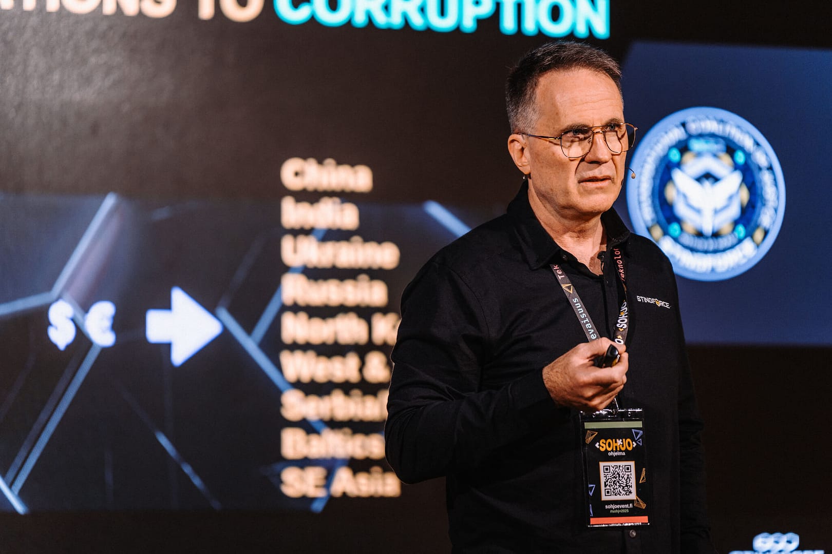A speaker in glasses and a black shirt, holding a clicker, stands on stage with a presentation slide in the background. The slide lists several countries and includes graphics of an arrow and currency symbols.