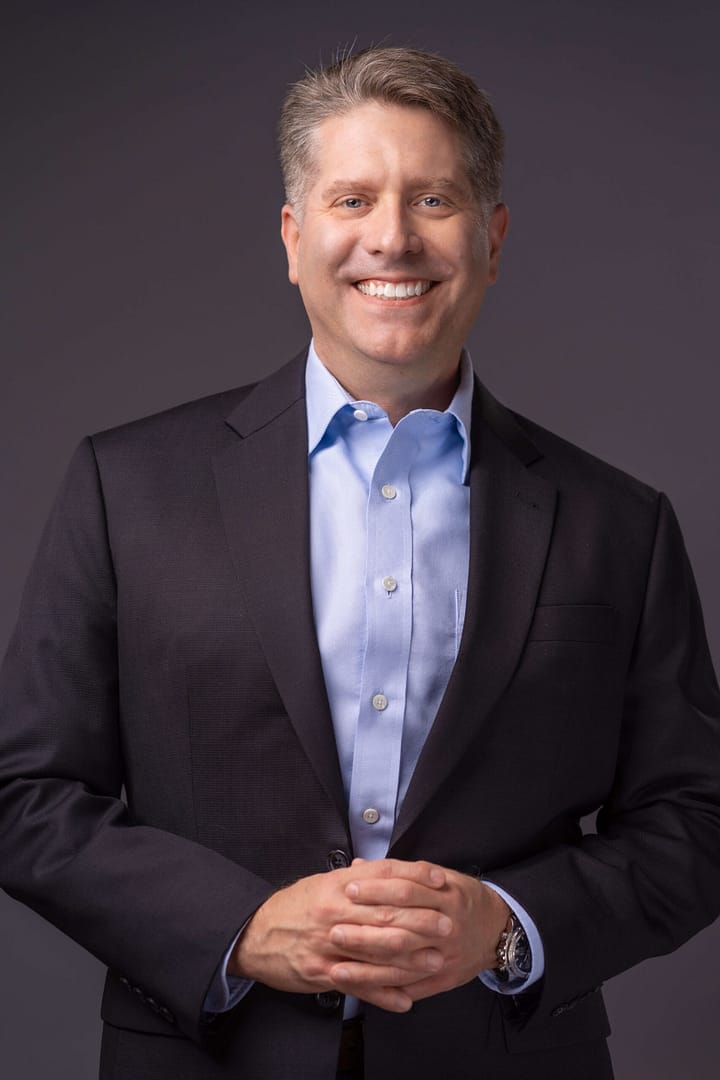 A person in a dark suit and light blue shirt smiles while standing against a gray background. They have short, light-colored hair and are clasping their hands in front.