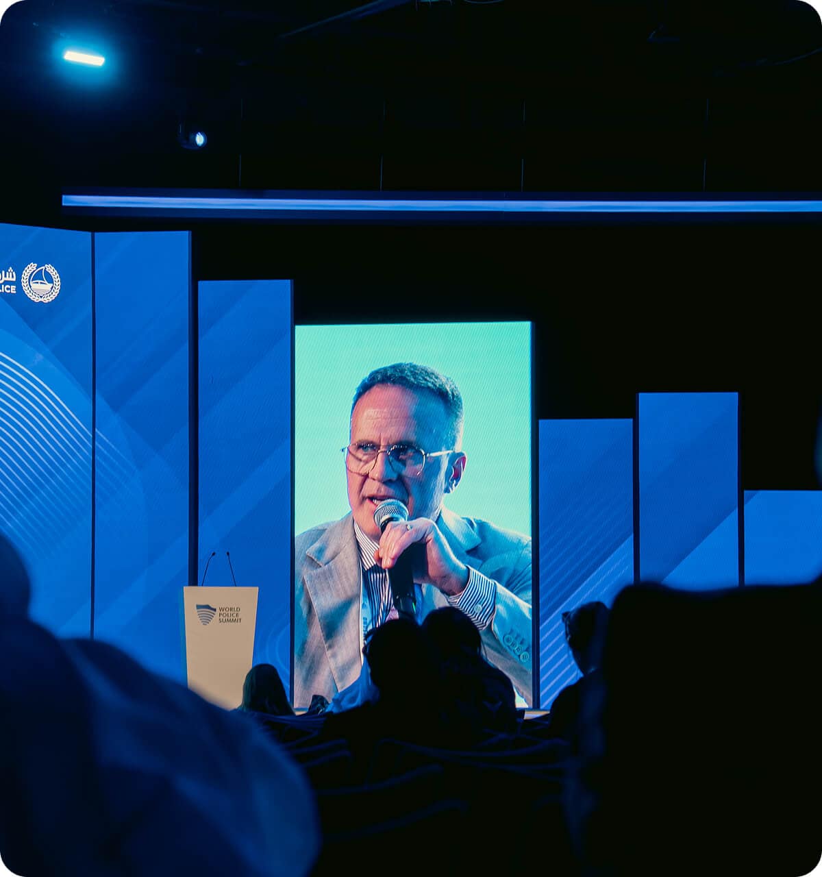 A man in a suit speaks into a microphone on stage at a conference, shown on a large vertical screen, with blue panels and an audience in the foreground.