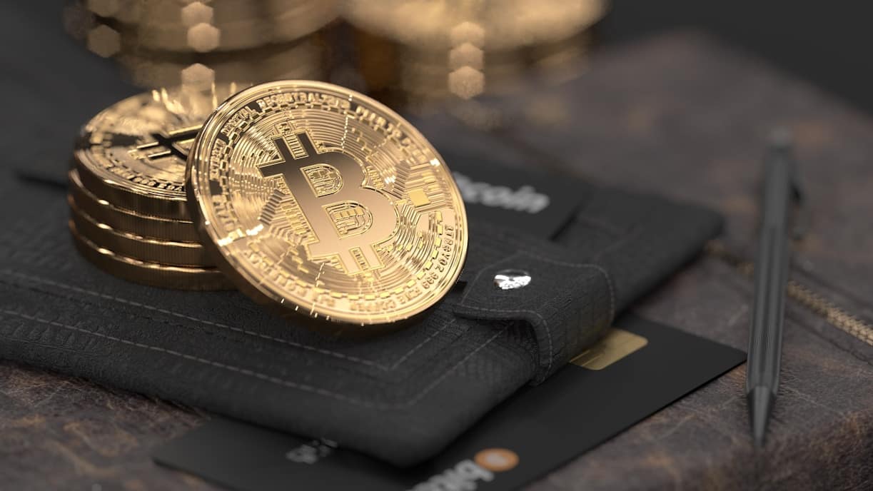 Two gold-colored Bitcoin coins rest on a black leather wallet, with a stack of coins, credit cards, and a pen nearby, symbolizing digital currency and finance.
