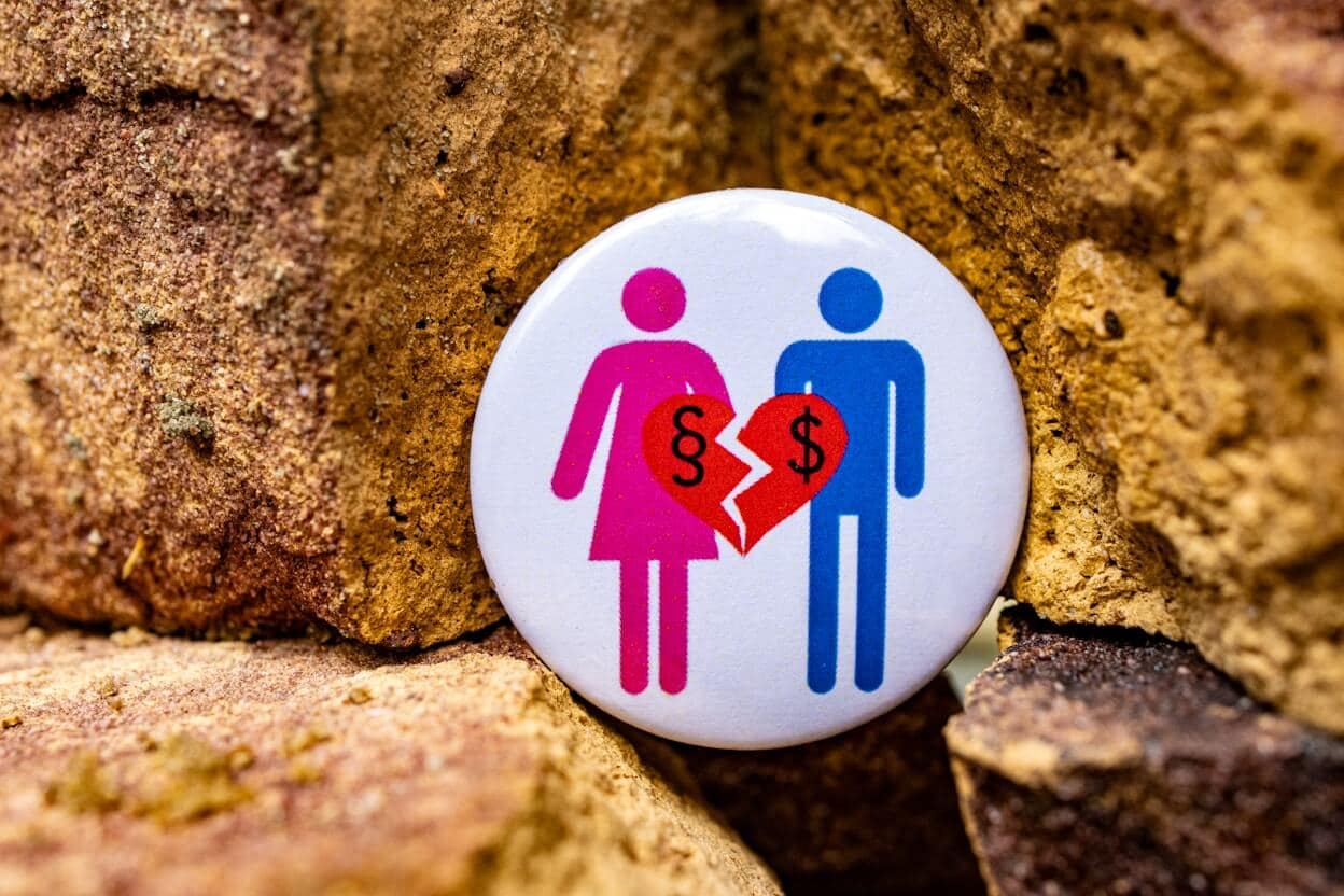 A button shows pink and blue icons of a woman and man holding a red broken heart with dollar signs on each half, symbolizing a financial split, against a background of rough brown bricks.