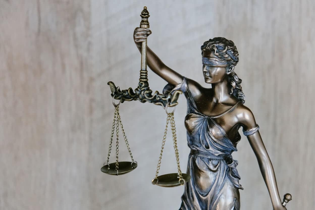A bronze statue of Lady Justice holding a balance scale in her left hand and a sword in her right hand. She is blindfolded, symbolizing impartiality. The background is blurred and neutral.