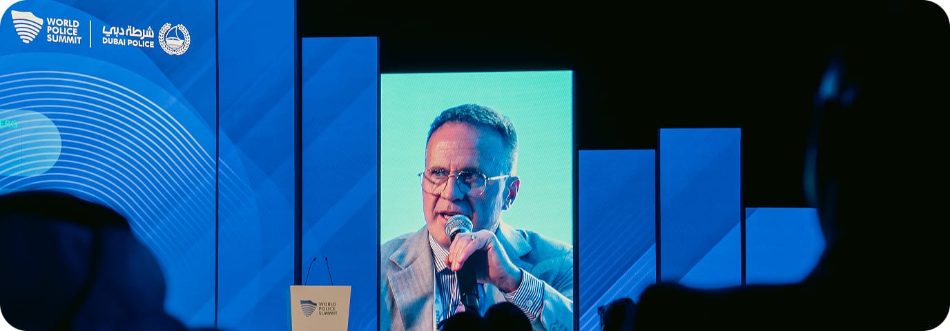 A man speaks into a microphone on stage at the World Police Summit, shown on a large blue screen with event logos and text in English and Arabic. Audience silhouettes are visible in the foreground.
