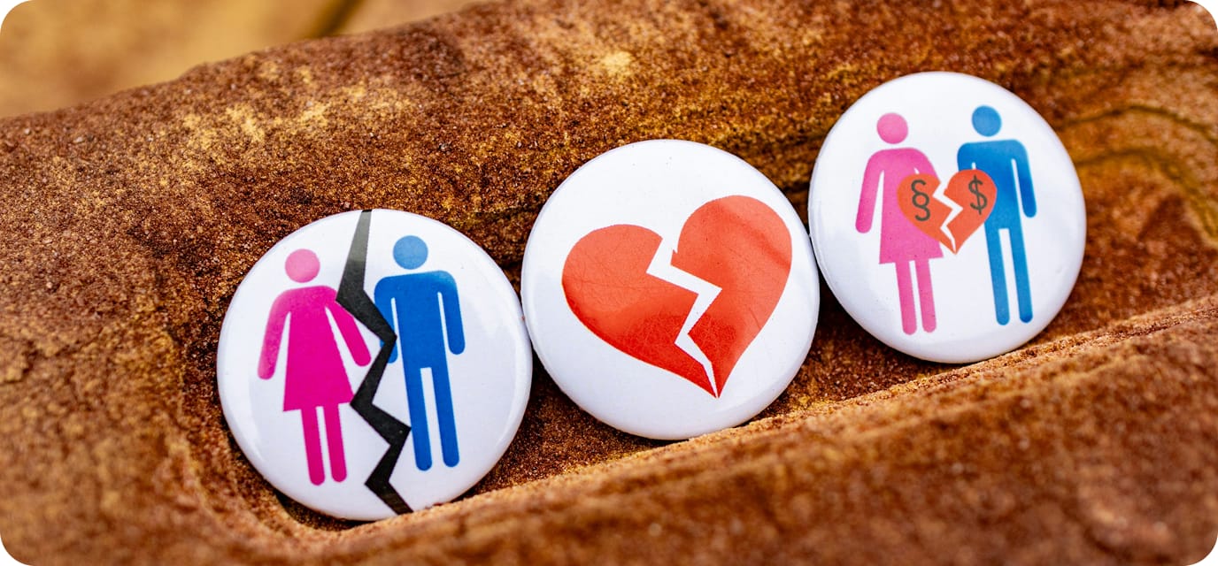 Three buttons on brown fabric: one with a cracked line splitting a male and female figure, one with a broken heart, and one with male and female figures holding a broken heart with dollar signs.