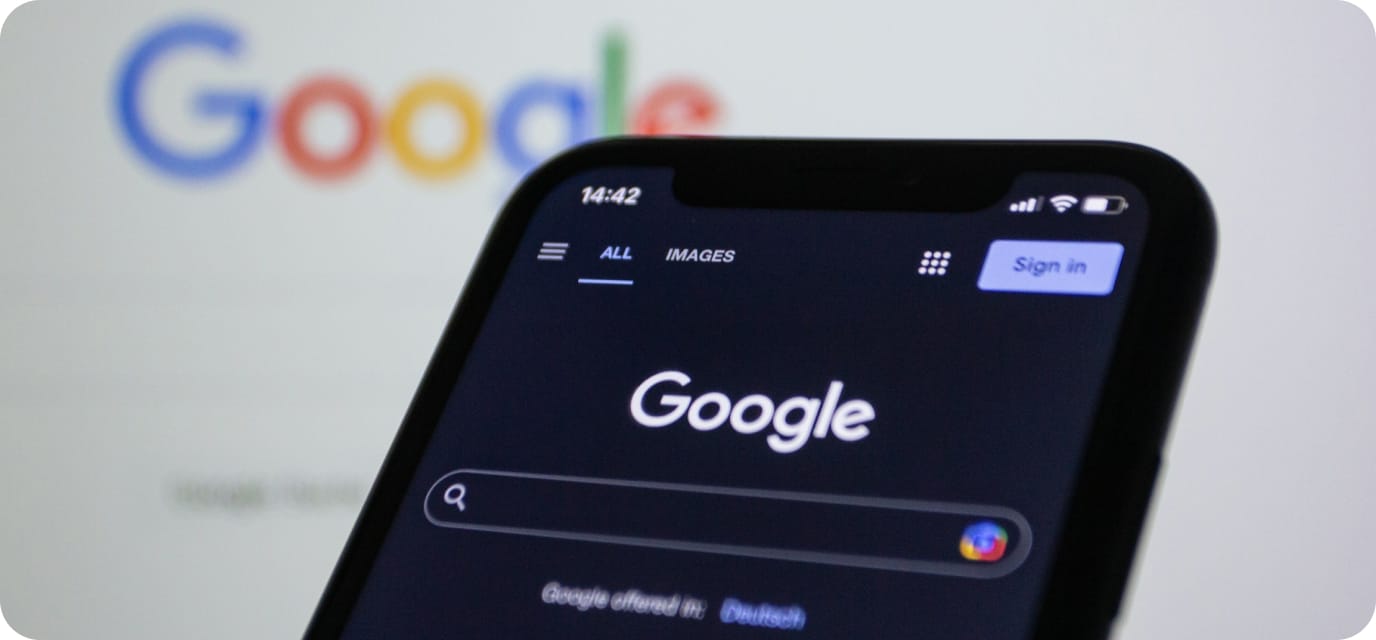 A smartphone displays the Google search homepage in dark mode, with the "Sign in" button visible. In the blurred background, the Google logo is shown on a computer screen.
