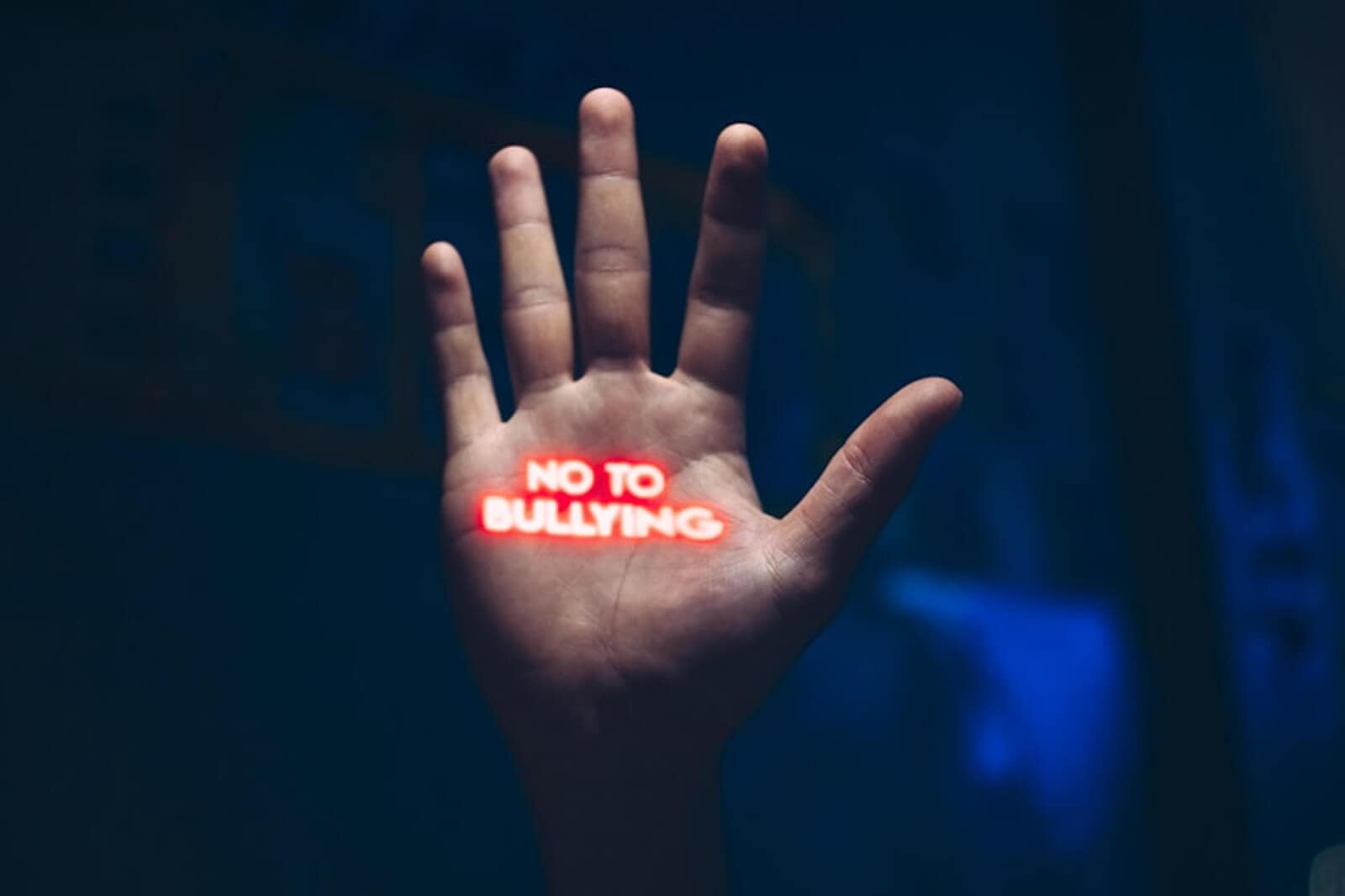 A hand held up with the words "NO TO BULLYING" in bold red letters projected onto the palm, symbolizing a stand against bullying. The background is dark and out of focus.