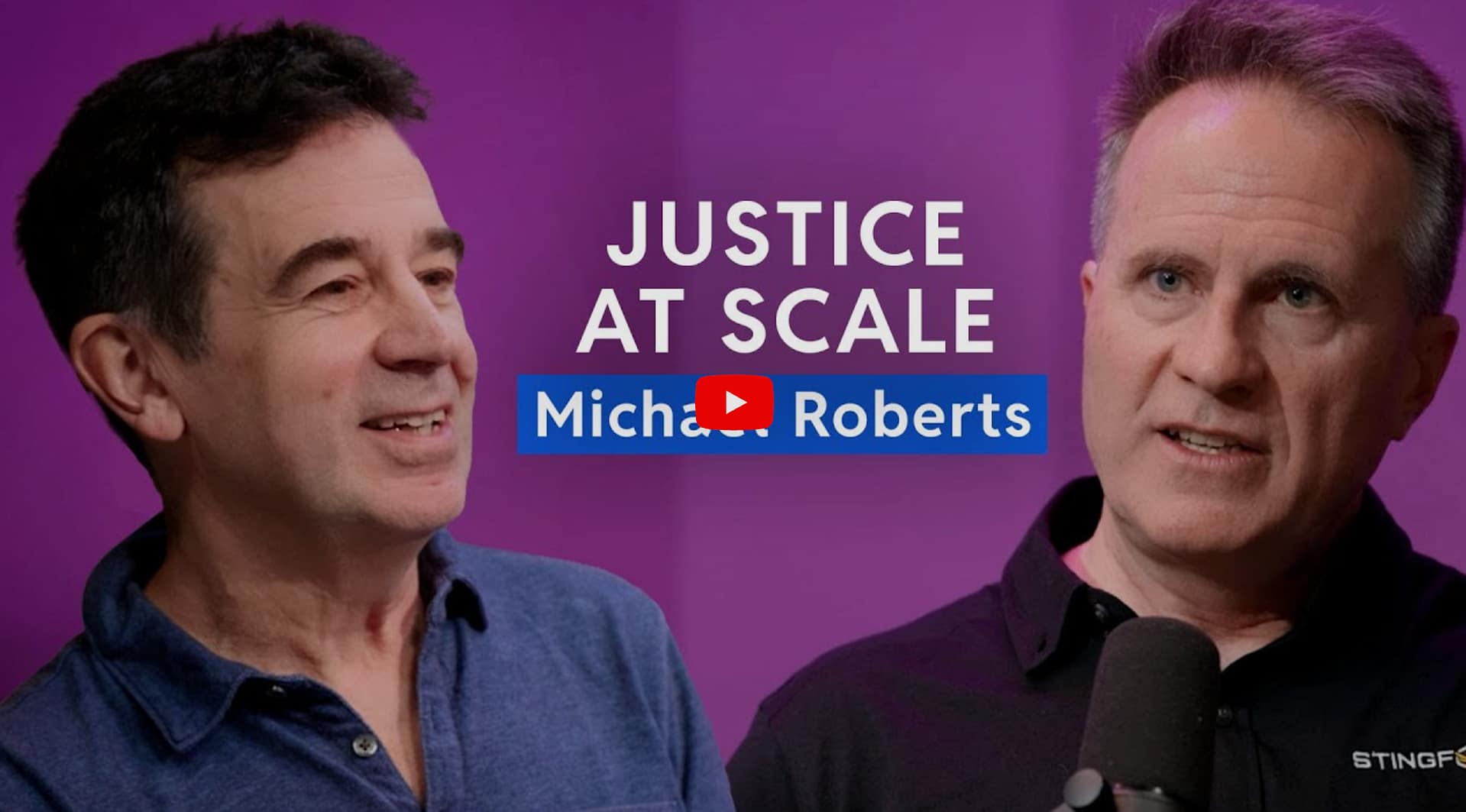 Two men are talking in front of a purple background. Text reads "JUSTICE AT SCALE Michael Roberts." One man faces forward with a microphone nearby, while the other looks toward him, both appearing engaged in conversation.