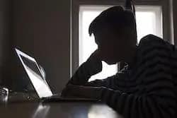 Silhouette of a person sitting at a desk, leaning forward and looking at a laptop screen, with light from a window in the background.