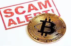 A gold Bitcoin coin lies on a white surface with the words "SCAM ALERT!" in bold red letters behind it, warning about cryptocurrency scams.