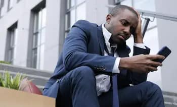 A man in a blue suit sits on outdoor steps, looking stressed or worried as he stares at his phone. His head rests on one hand, and office buildings are visible in the background.