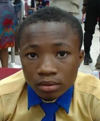 A boy with short hair wearing a yellow shirt and a blue tie looks directly at the camera. He is indoors, with people and colorful clothing visible in the background.