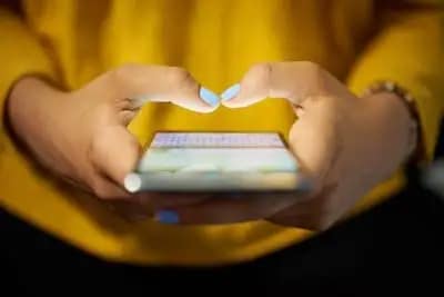 Close-up of a person with light blue nail polish typing on a smartphone, wearing a yellow top. The phone screen is slightly blurred and the person's hands are in focus.