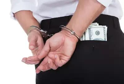A person in handcuffs with their hands behind their back, wearing dress pants with cash sticking out of the back pocket.