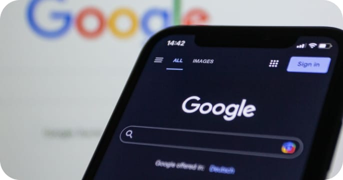 A smartphone displays the Google search homepage in dark mode, with the Google website also visible blurred in the background on a larger screen.